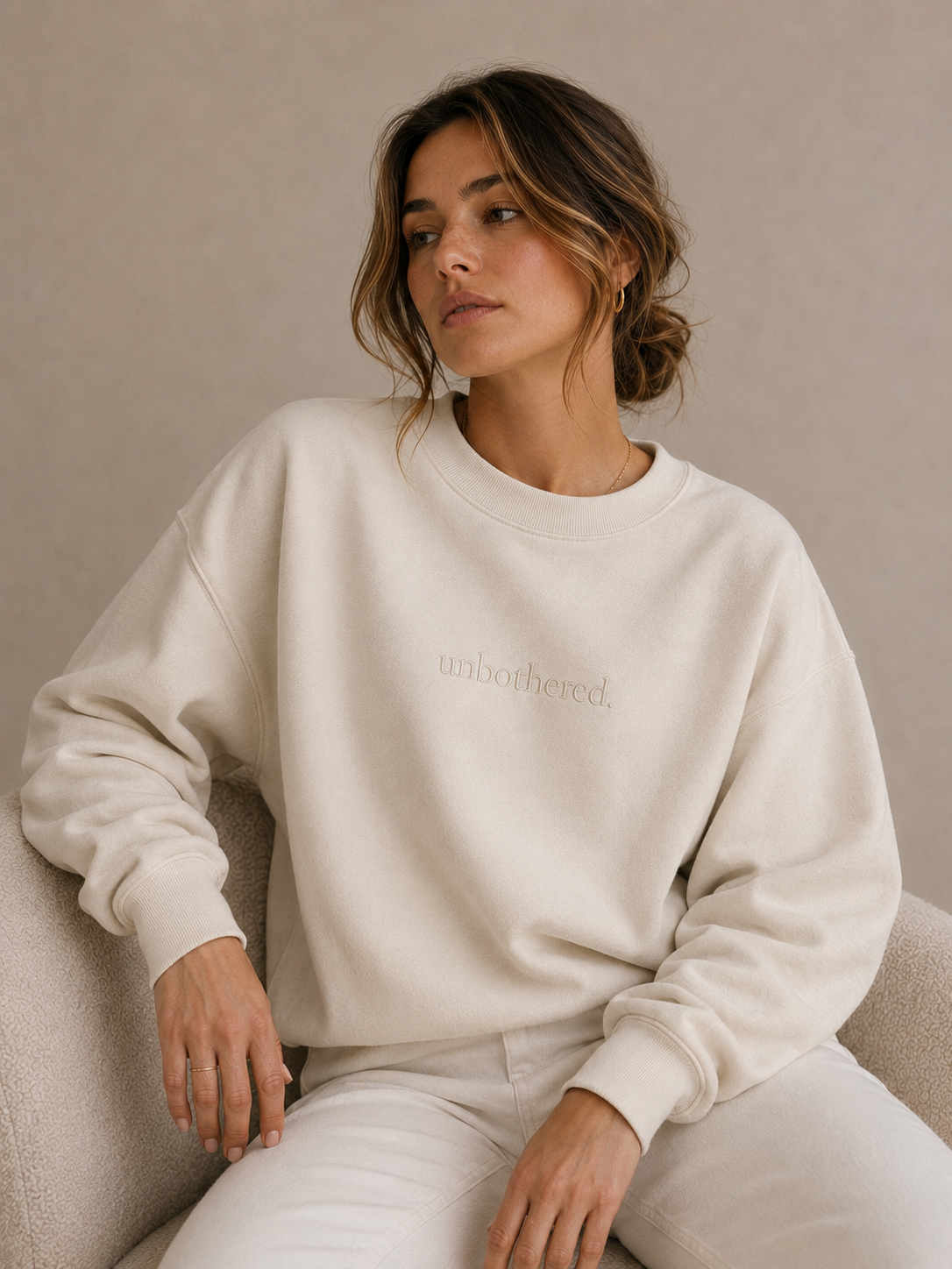 Woman wearing a cream sweatshirt with 'unbothered' text, sitting on a beige couch.